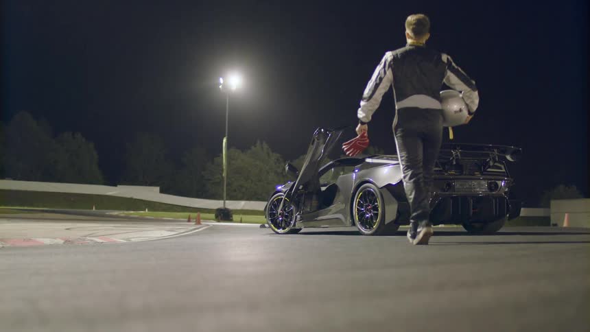 A race car driver carries his helmet as he walks towards the open drivers seat door of a car parked next to a race track at night