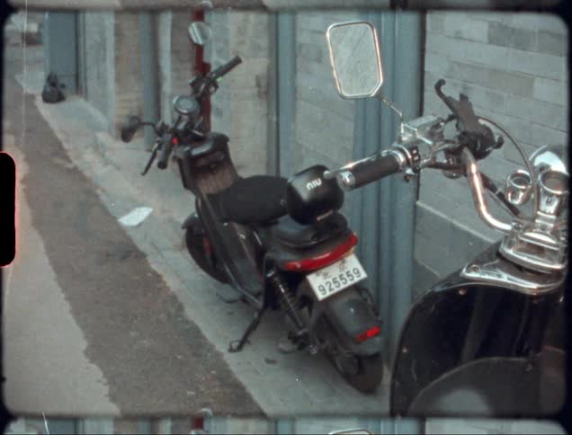 Two mopeds sit on side street in front of a wall made of grey brick with trash too the side
