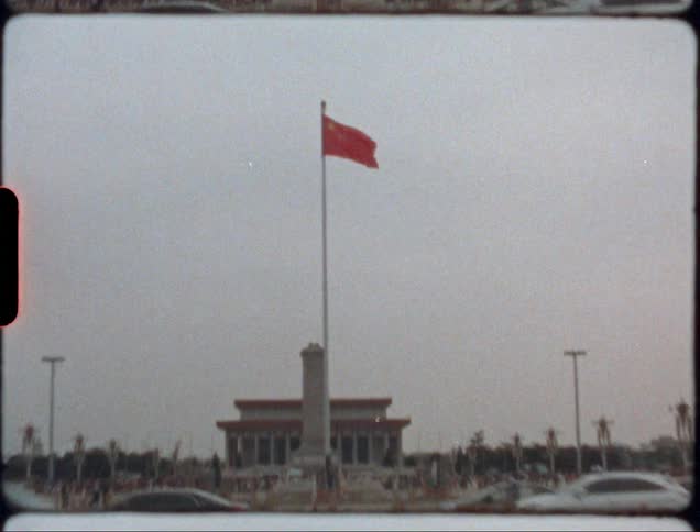 Zoom in to a red flag on top of a flag pole and out to show cars driving on a street