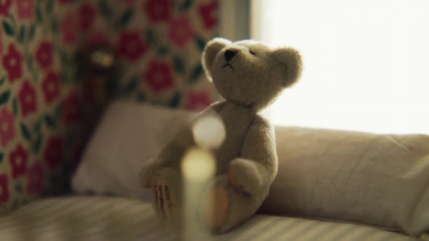 Small brown teddy bear on a cushion at the head of a made bed