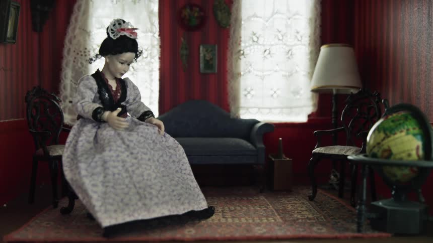 Hand placing a tiny doll onto a chair in a miniature living in a doll house