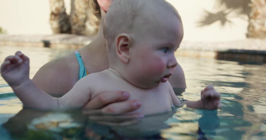 A woman holding a baby in a pool with trees and a wall in the background