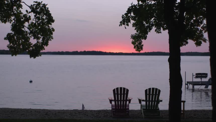 A pair of a lounge chairs sitting side by side on the shore of a lake at sunset