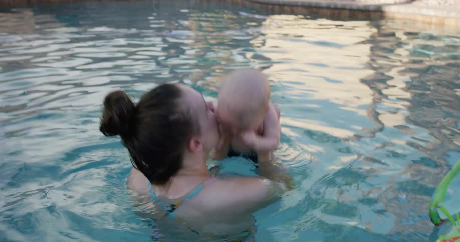 A mother is holding and kissing her baby while standing in a pool outdoors during the day