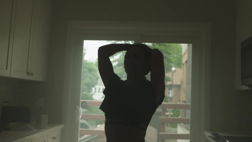 A person stands in an unlit kitchen near a doorways with hands held up to the head shifting back and forth on feet