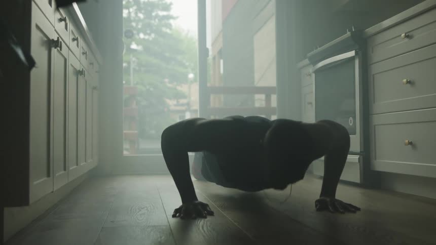 A man does push ups on the wooden floor of a small home kitchen illuminated solely by sunlight from a glass door