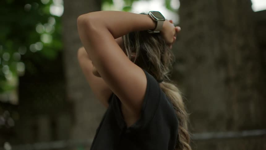 A teenage girl stands in a neighborhood with her hands behind her head and looks around