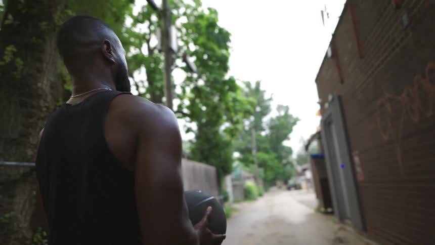 A man tosses a ball to a woman as they exercise and lunge down an alley outside during the day
