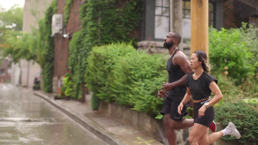 A young woman and man in athletics clothing run through a neighborhood in the rain during the day