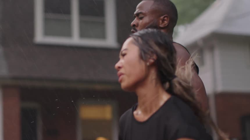 A young woman runs through the rain along a street in a neighborhood alongside a man