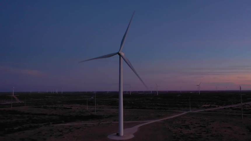A white wind turbine stands in a line with other turbines with the sky changing colors at sunset