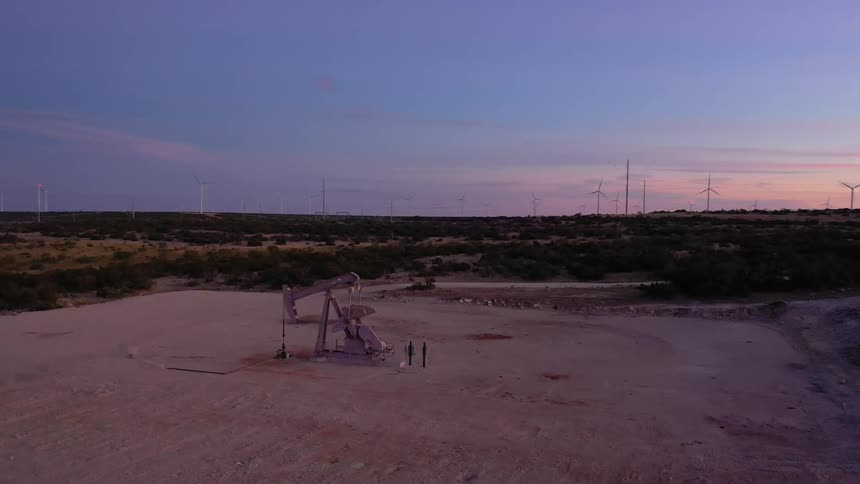 The sun sets on a remote area filled with oil derricks pumping oil and windmills spread all over the sandy brush filled land