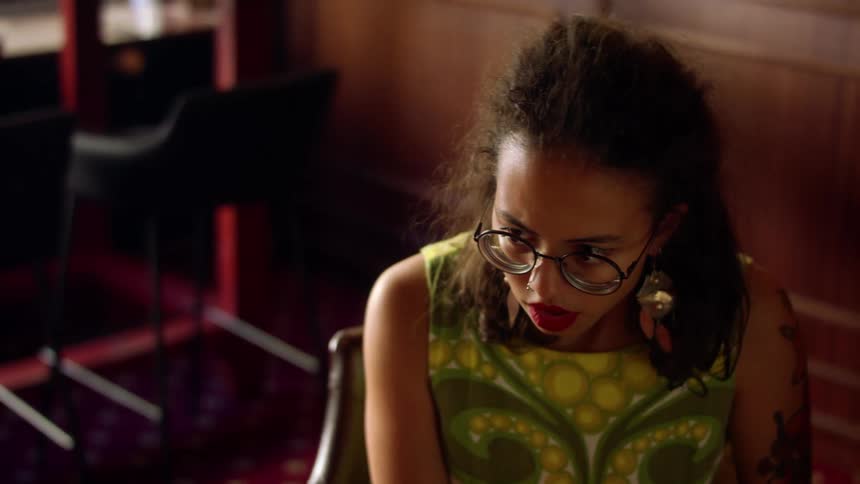 A young woman sitting in a chair indoors wipes her cheek with the back of her hand