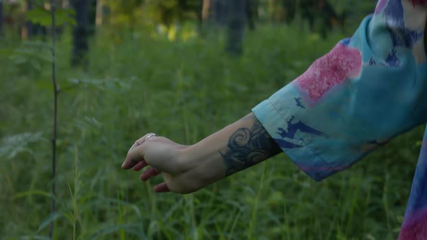 Person with tattoos moves their hand in flowing gesture while standing in forest