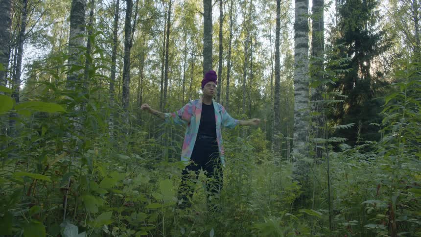 A woman wearing a turban and a colorful jacket dances in a forest