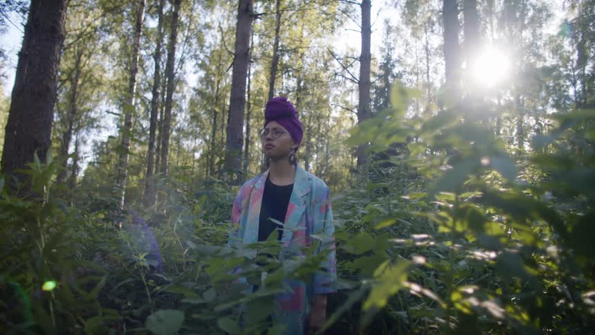 A woman with glasses standing in the woods with trees the sky and clouds in the background