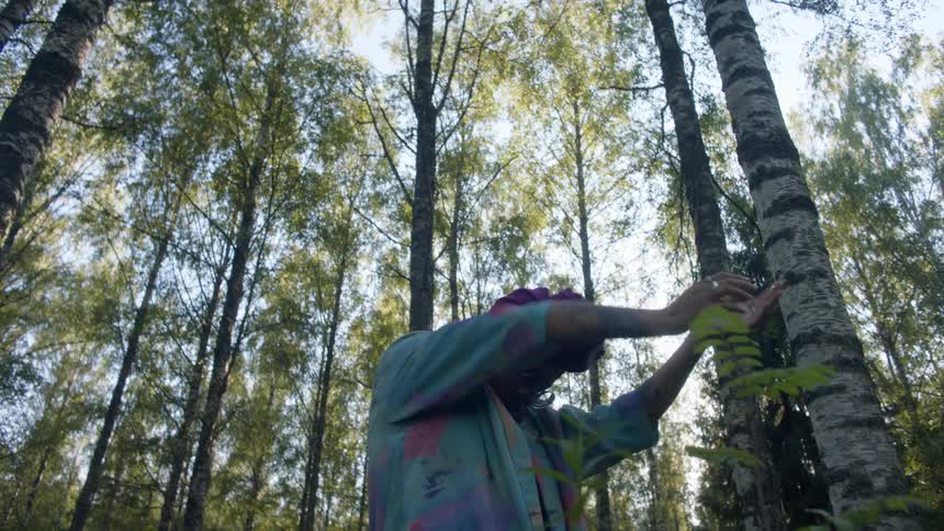 A woman with glasses dancing in the forest with the sky and clouds in the background