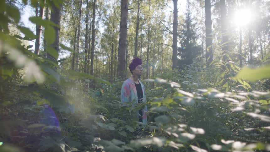 Woman wearing turban and blue coat stands in forest clearing under sunshine