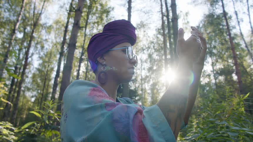 A young woman stands in a forest and holds her hands up in front of her face and examines them