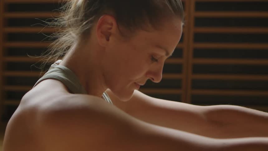 A woman lies on her back stretching and then sitting up on a wooden floor inside exercising during the day