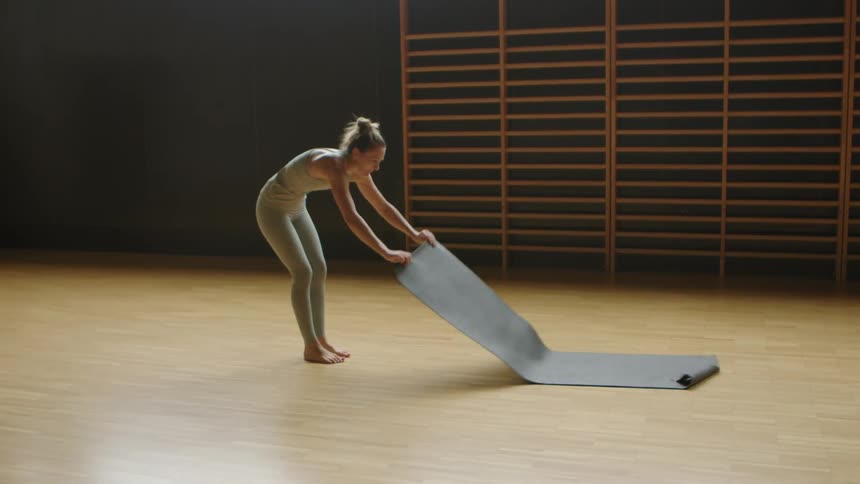 A woman unrolls and stands on a yoga mat in a gymnasium during the day