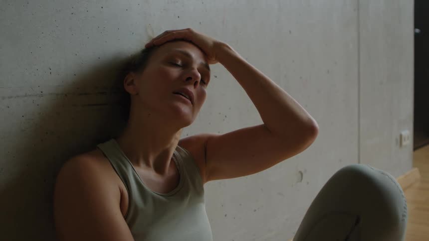 A woman stands next to a wall and slides down to sit down and catch her breath indoors after exercising