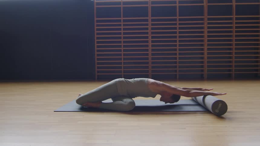 A woman is seated on the floor and is performing yoga poses and using a foam roller