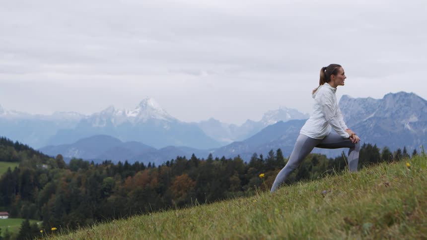 A woman stretches her leg on an incline of a hill outside with mountains in the background with overcast weather