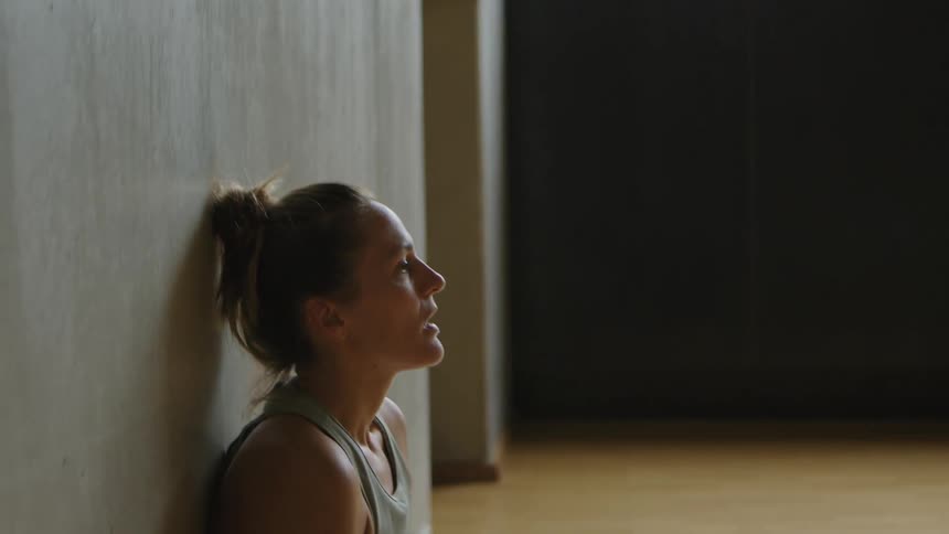 A young woman catching her breath after exercising leans against a wall and slides down to sit on the floor