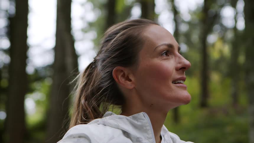 A woman working out outdoors stops in a forest to rest and runs her hands across the top of her head
