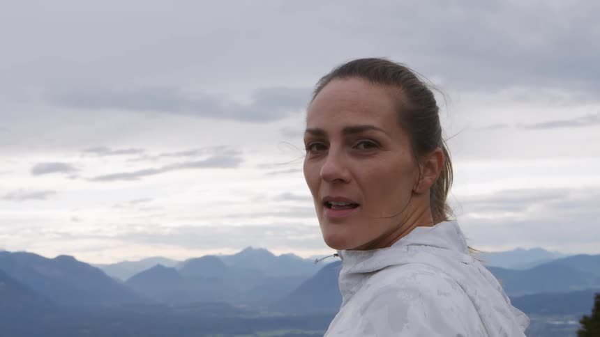 Woman with a ponytail and a white zip up hoodie faces the camera with eye contact on a cloudy day and with mountains in the background