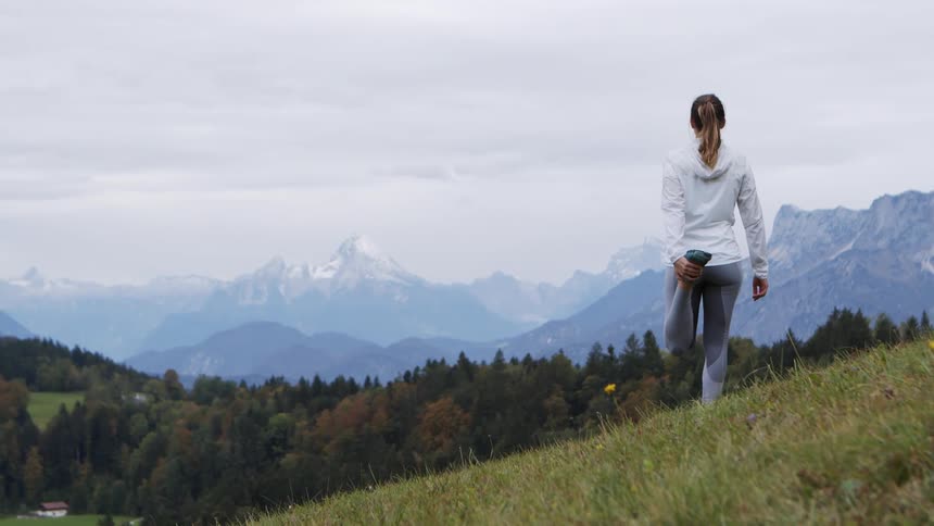 A woman stands and stretches atop a hill while facing mountains