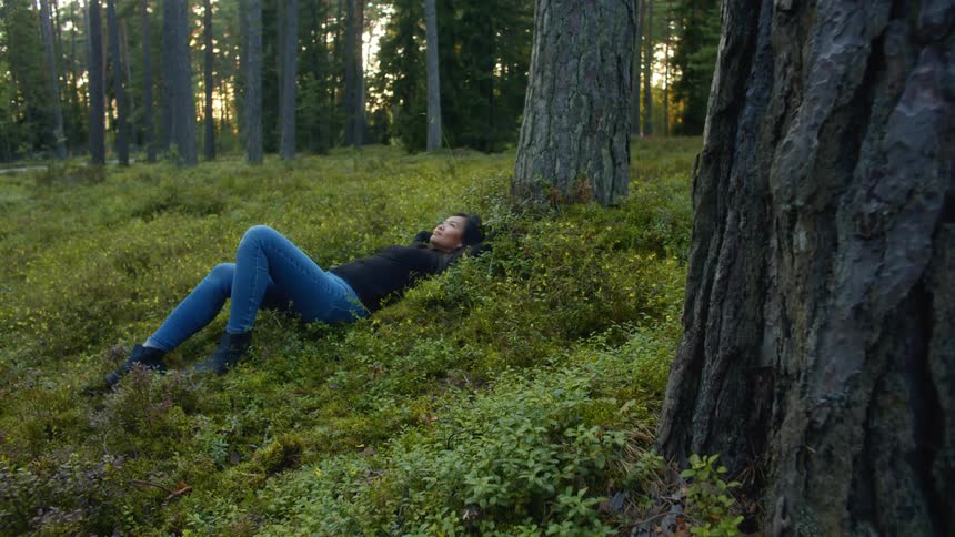 A woman lies on her back in grass in the forest outside at dusk