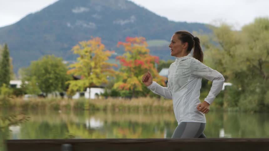 A woman runs outdoors along a river with trees and mountains during the day
