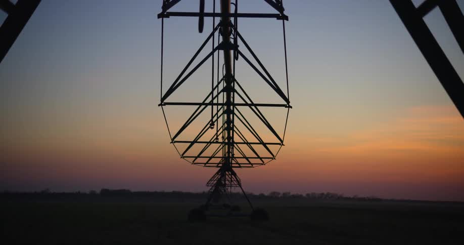 The sun sets over a field of crops where tall farm equipment sits