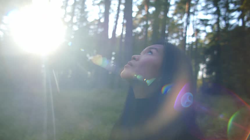 Woman with dark straight hair and a black sweater looks up at the sky as the sun shines through trees in the forest