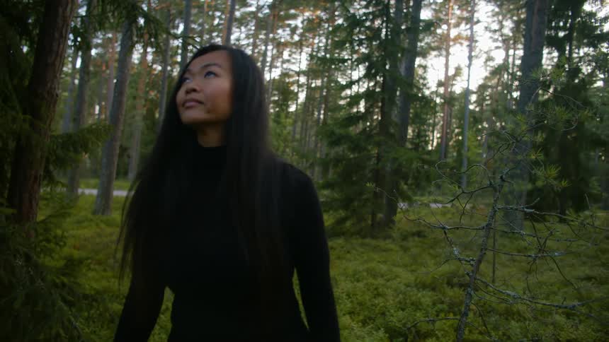 An asian woman examines her surroundings as she walks through a woodland during the day