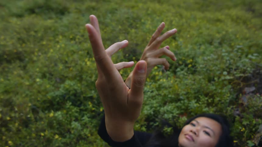 A woman twirls her hands as she lays on her back among the vegetation in a field in the evening