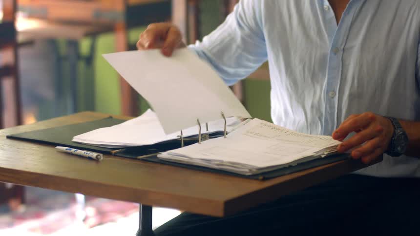 A man sits at a small desk flipping through papers in a binder