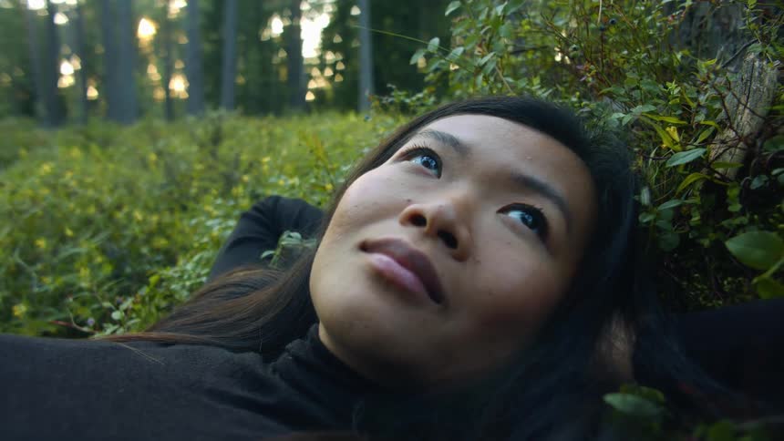 Asian woman with straight hair and a black sweater lies down on the grass in the forest