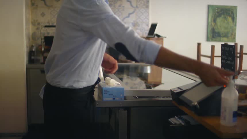 A man stands behind the corner of his restaurant and walks over to the register to use hand sanitizer