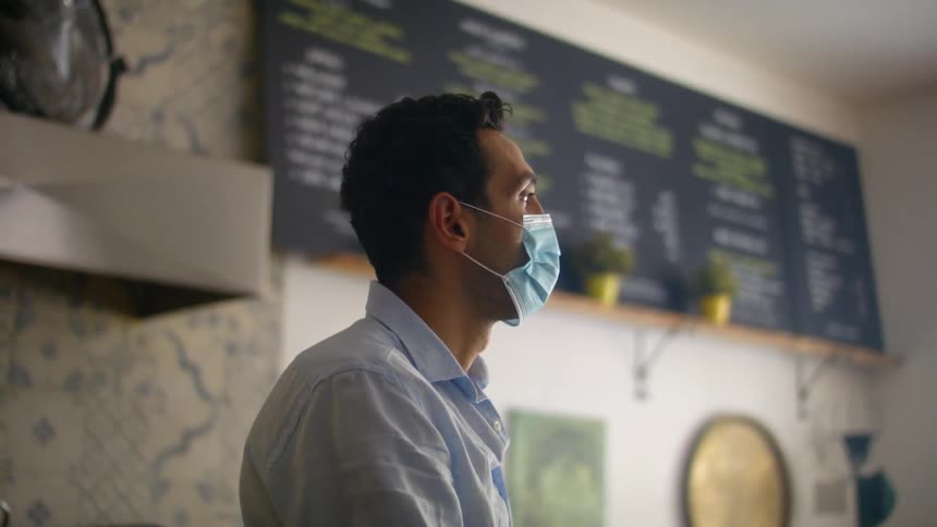 Barista with a blue mask and a shirt stands in a coffee shop