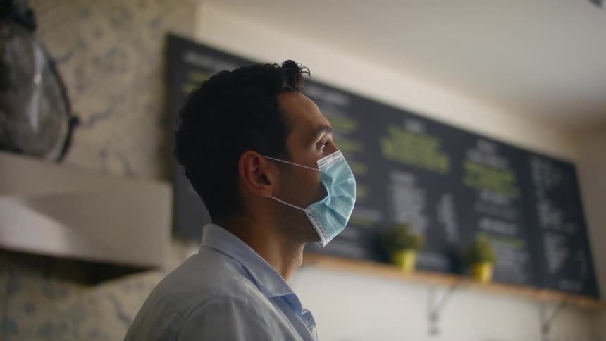 A man in a medical mask closes his eyes for a moment as he stands contemplatively in a restaurant