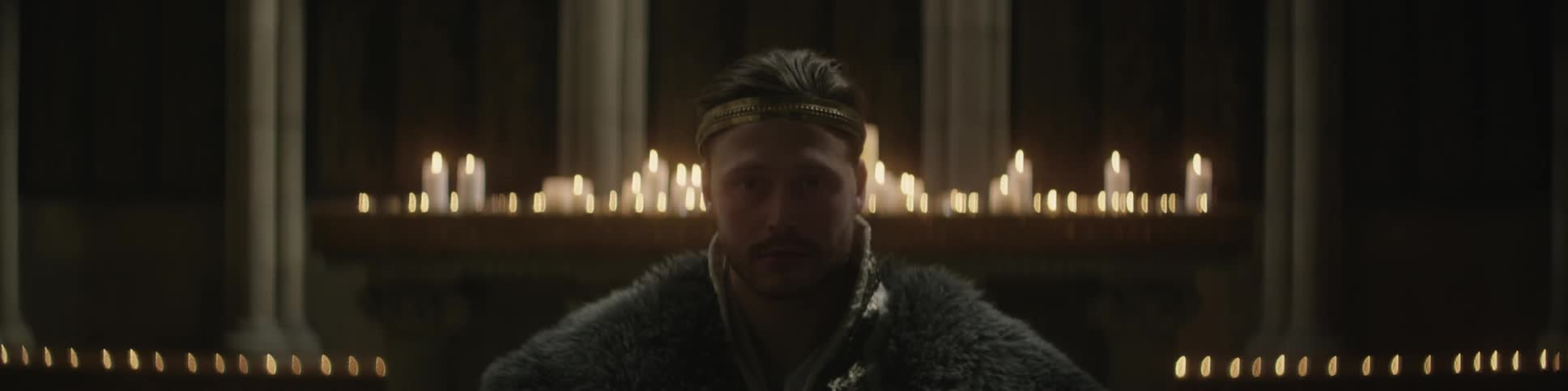 Man wearing fur and crown sitting in front of burning candles