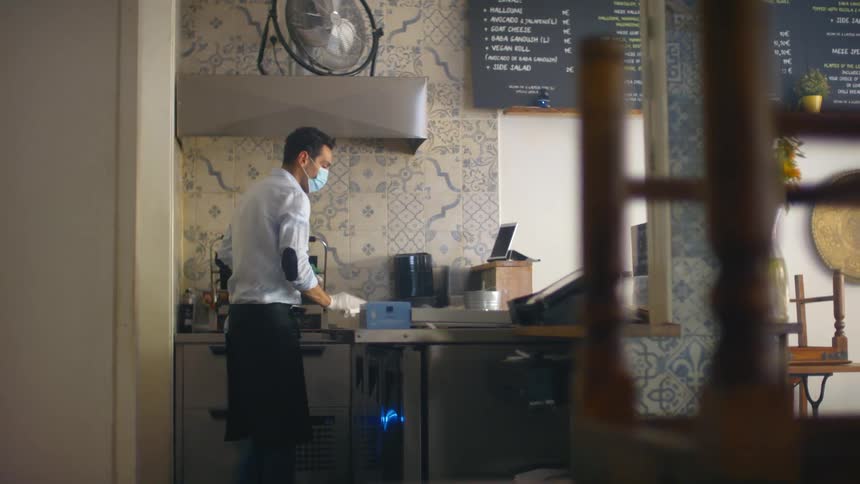 A man in an apron and mask wipes a counter with a washcloth in an unopened restaurant during the day