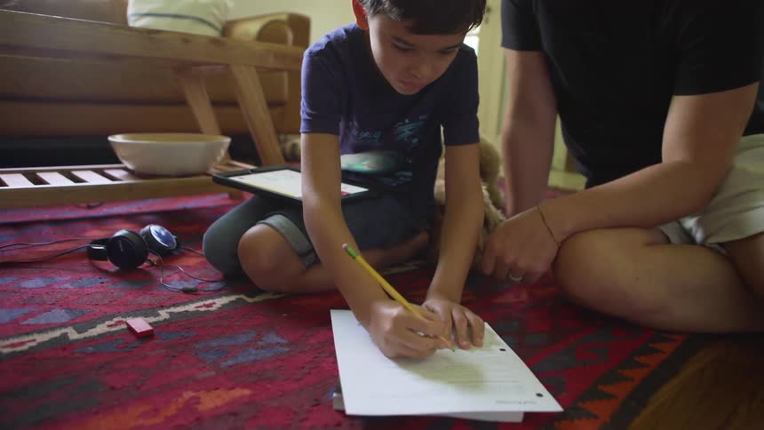 A man kneels on the floor beside his son writing on a piece of paper