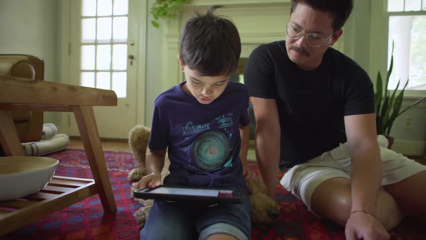 A father and son kneel on the floor looking at a tablet
