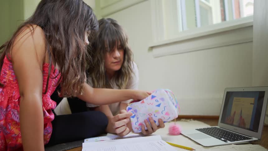 Mother holds bag of pencils for daughter to pick one in front of a laptop showing instructions on a table in front of a work book