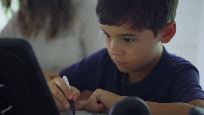 A boy copies text from a monitor onto a white board while sitting at a desk with his mother