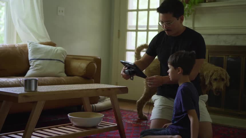 Father and son sitting in a living room setting up a tablet on a coffee table while dog walks around them